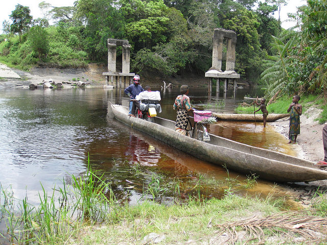 A dugout ferry takes motorbikes across the Kasuku River. The ancient bridge pillars in the background attest to colonial ambition – never realised.